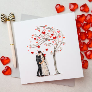 Handmade  quilling greeting card with bride and groom standing under a heart tree on a table with red hearts. 